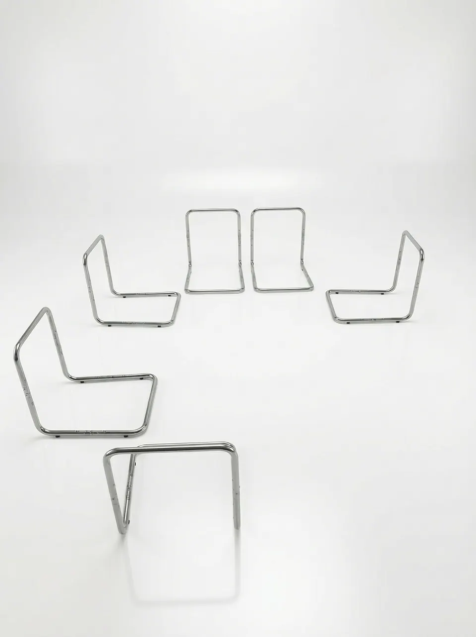 Six chrome tubular L-shaped seat frames arranged in a loose arc on a white studio floor with soft reflections