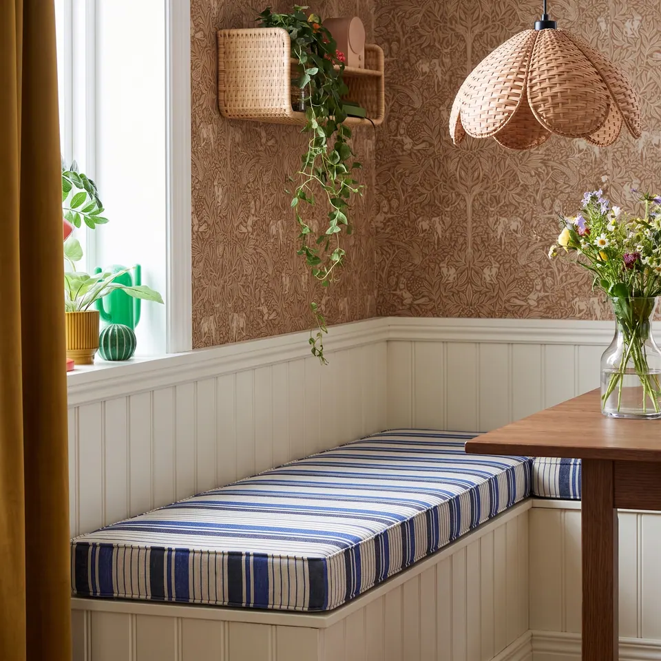 Blue and cream striped box cushion on a white beadboard corner dining bench with woodland wallpaper and rattan pendant