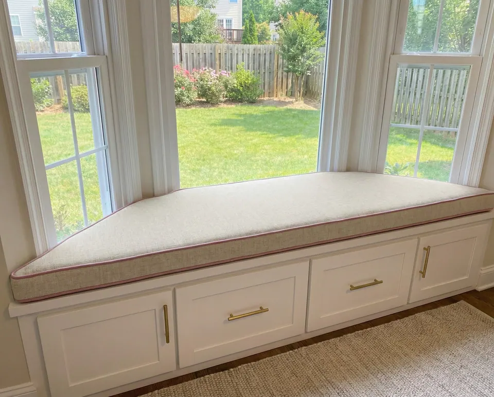 Oatmeal linen trapezoid cushion with blush pink piping on a white bay window bench with brass pulls, garden and fence beyond
