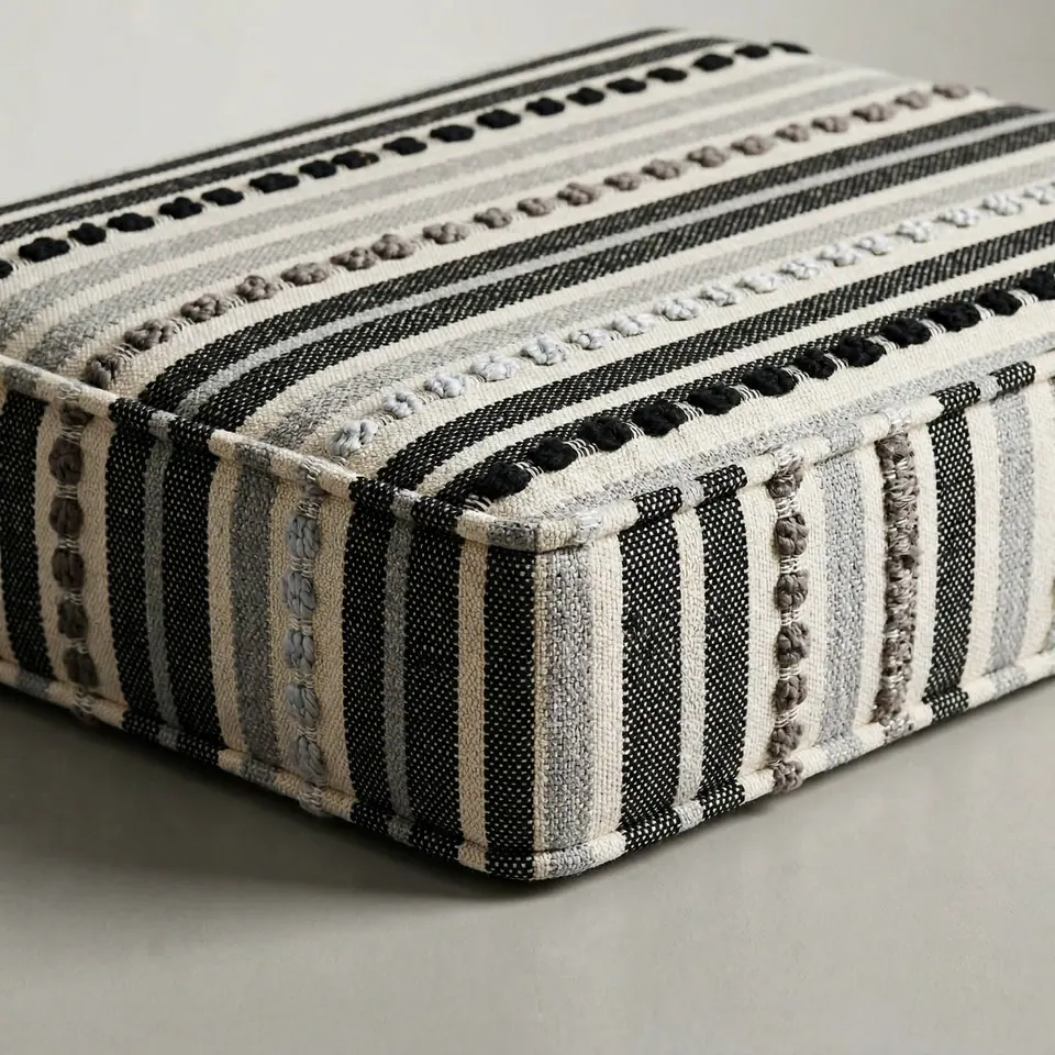 Corner of a box cushion in black, gray and cream woven stripes with raised knot rows and self piping on a gray backdrop