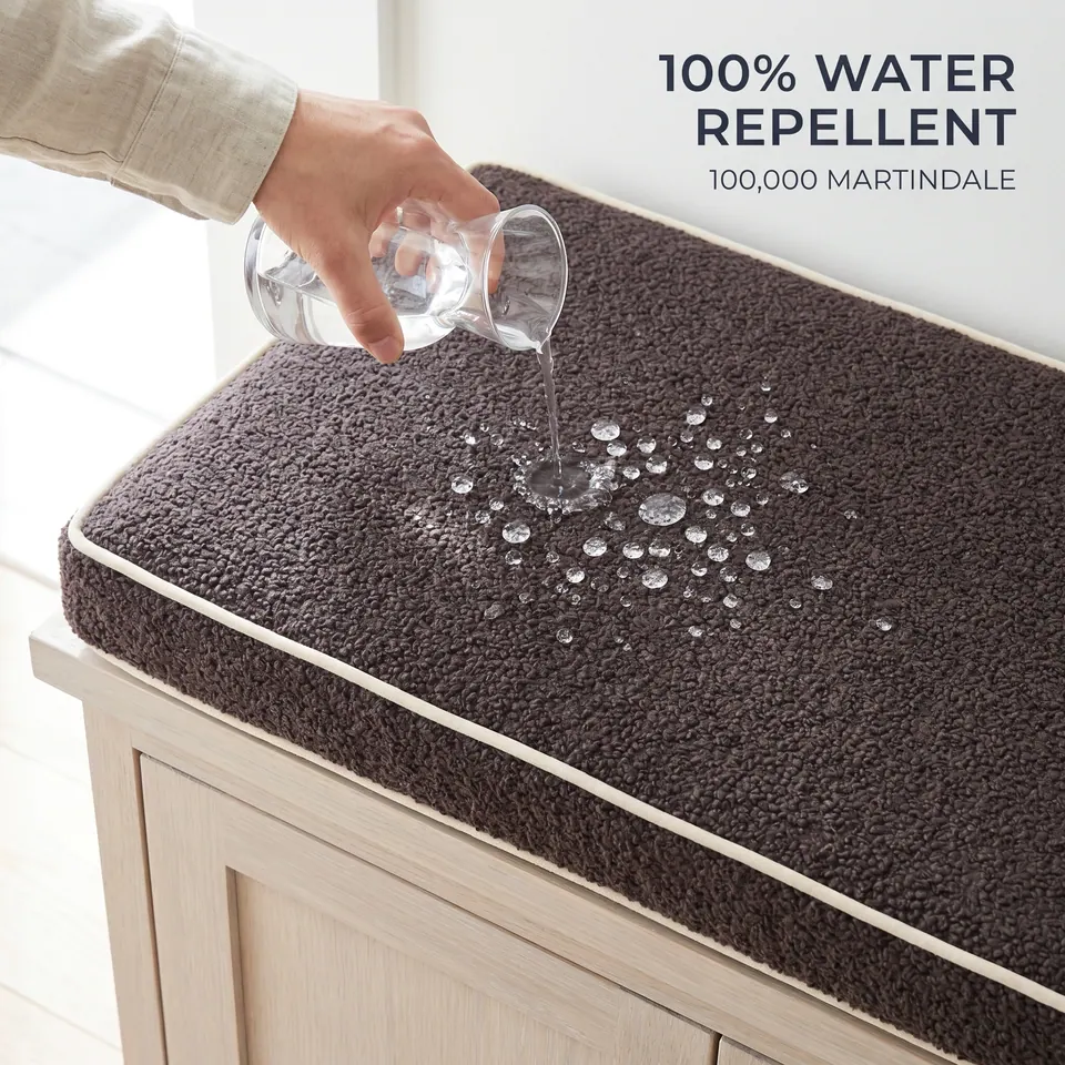 Water poured onto a charcoal brown bouclé bench cushion with cream piping forms beads on the surface, pale oak cabinet below