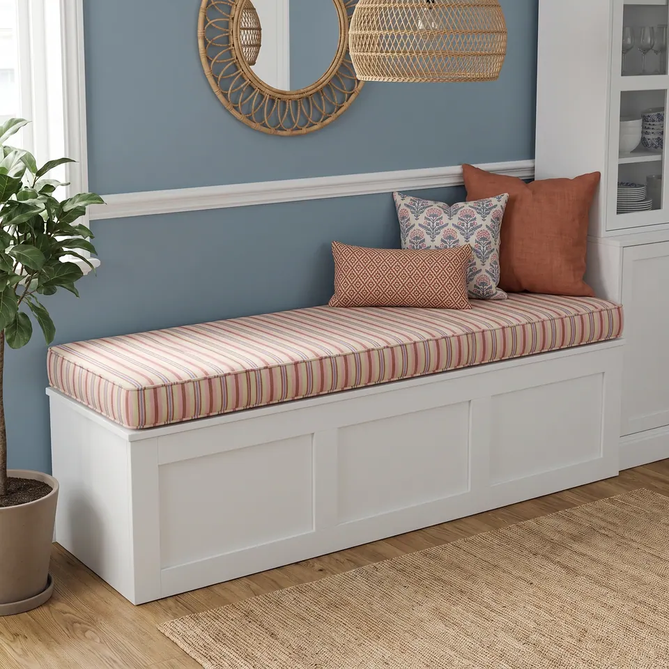 Cream box cushion striped in coral, pink and sage on a white shaker storage bench against a dusty blue wall with rattan decor