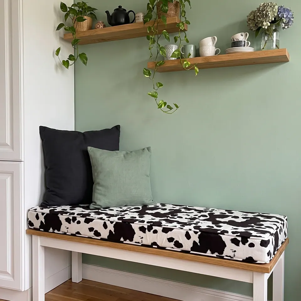 Black and white cow-print bench cushion on an oak-topped bench against a sage wall under floating shelves of cups