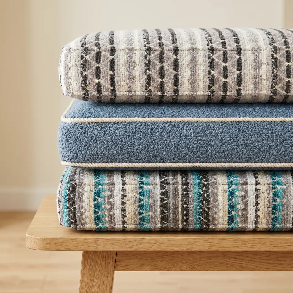 Dusty blue bouclé cushion with cream piping stacked between gray and teal woven-stripe box cushions on oak