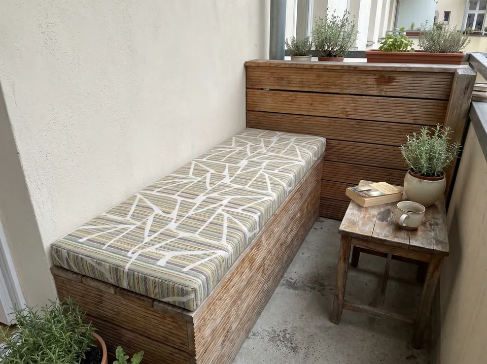 Pastel multi-stripe outdoor cushion with white branch lines on a slatted storage bench on a narrow city balcony