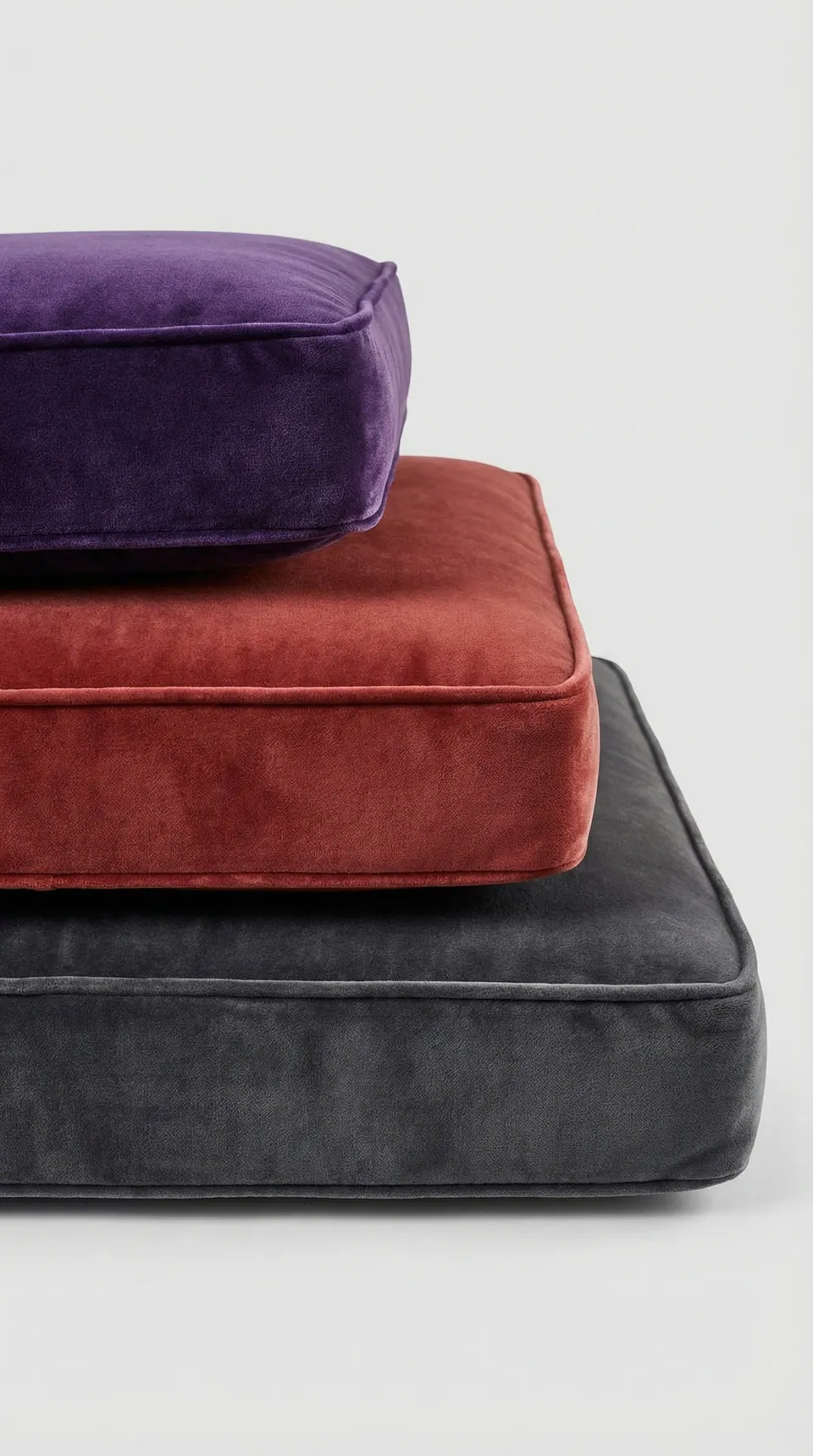 Three stacked velvet box cushions in violet, rust red and charcoal gray with matching piping on a pale gray backdrop