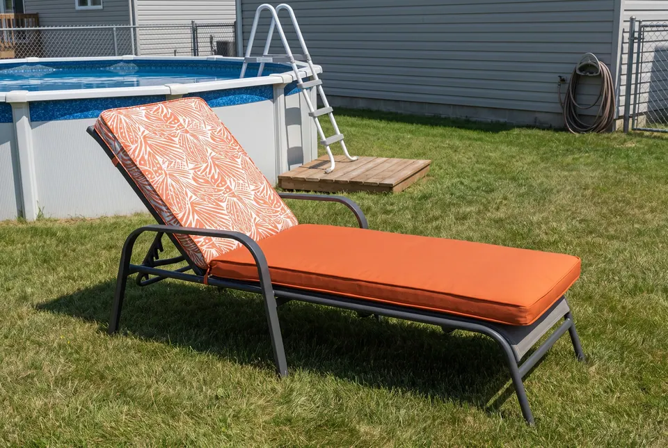 Two-fabric cushion with orange and cream leaf-print back and solid orange seat on a steel lounger by an above-ground pool