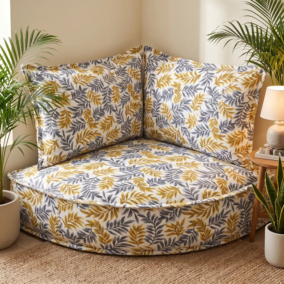 Gray and gold leaf-print corner floor cushion with backrest pillows between potted palms and a lit table lamp