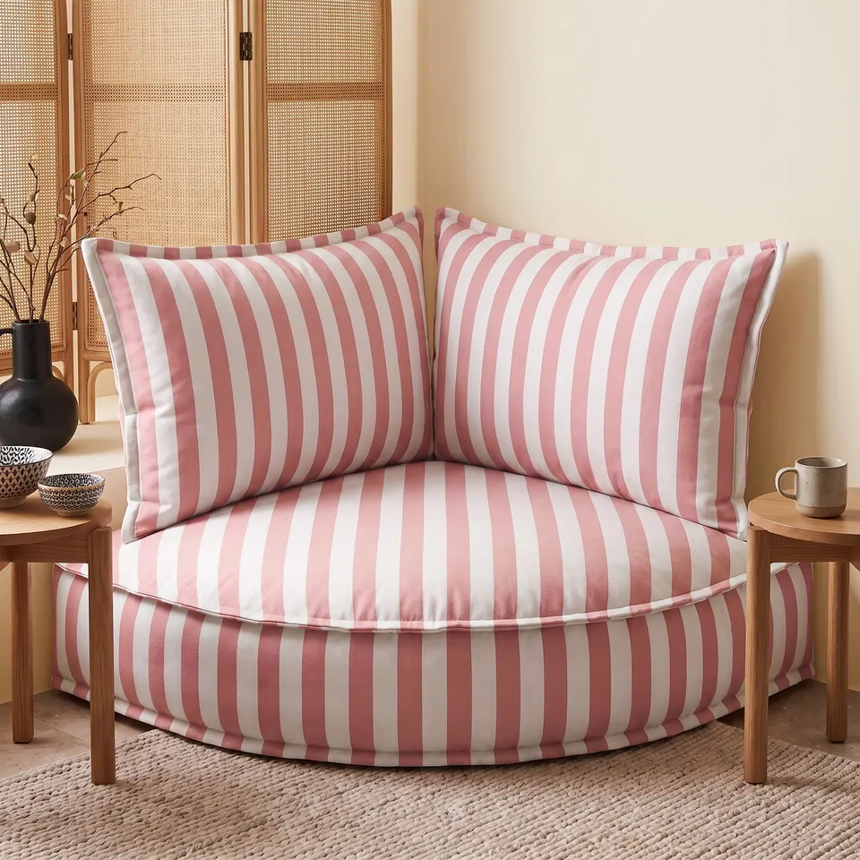 Rose pink and white striped corner floor pouf with backrest pillows between two oak side tables, rattan screen behind