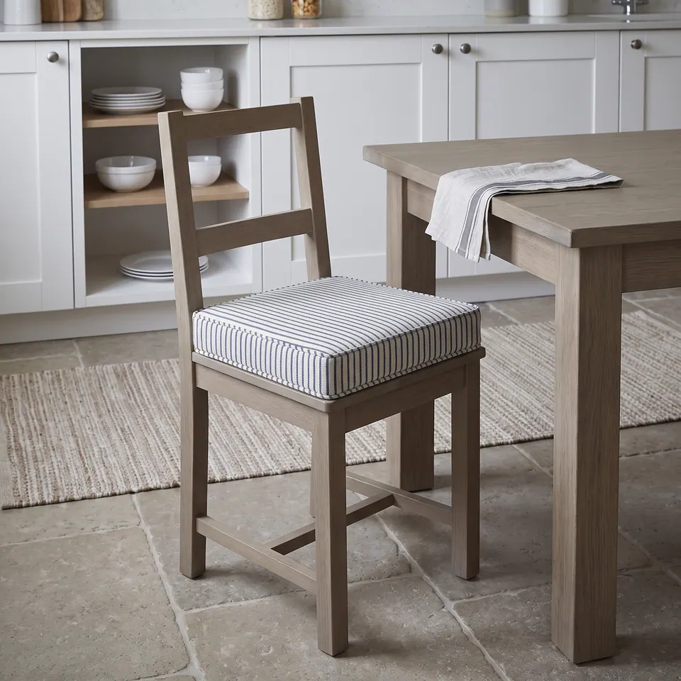Indigo ticking stripe box cushion on a weathered oak ladder-back chair in a white kitchen with limestone floor