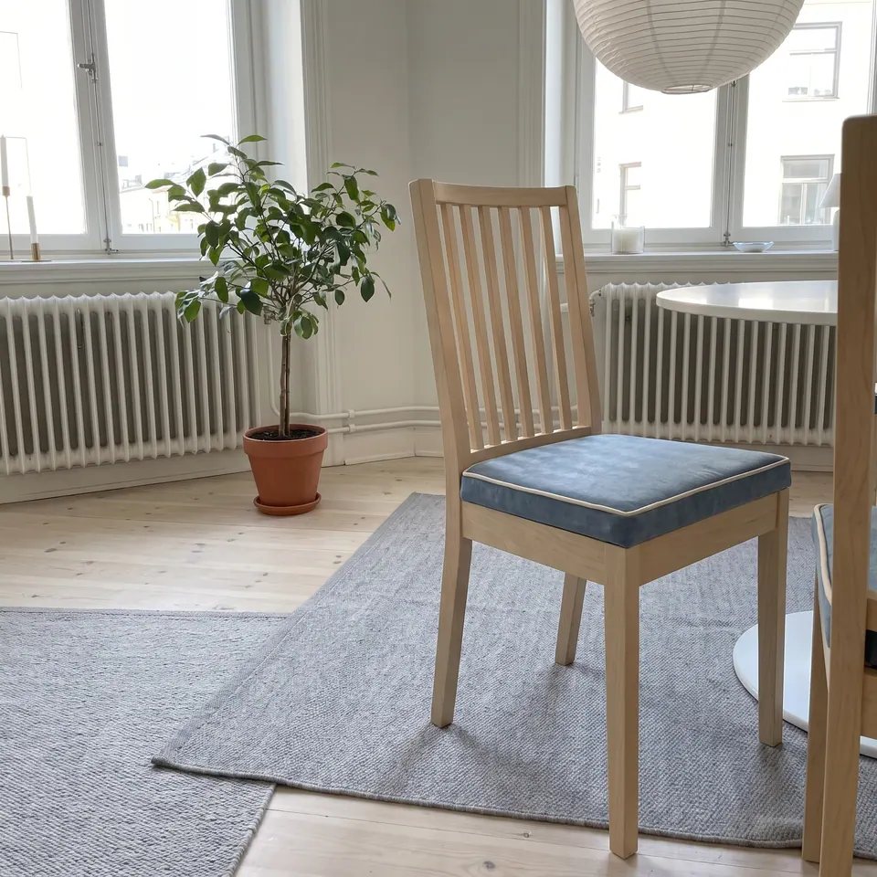 Steel-blue linen-look seat pad with cream piping on a pale birch slat-back chair, gray rug, potted fig and white radiators