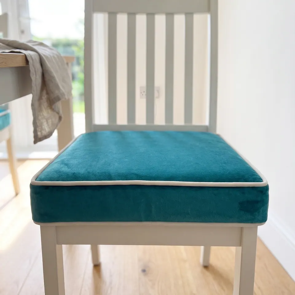 Straight-on view of a teal velvet box cushion with off-white piping on a white chair, showing its deep front gusset