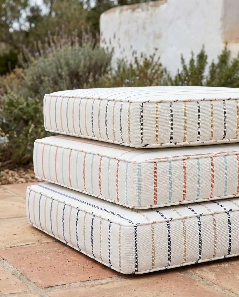 Three cream box cushions with navy, coral and mustard stripes stacked on terracotta tiles in a courtyard with rosemary