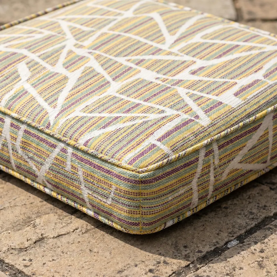 Close-up of a box cushion corner in mustard, plum and green woven stripes with a white branch pattern and piping on stone