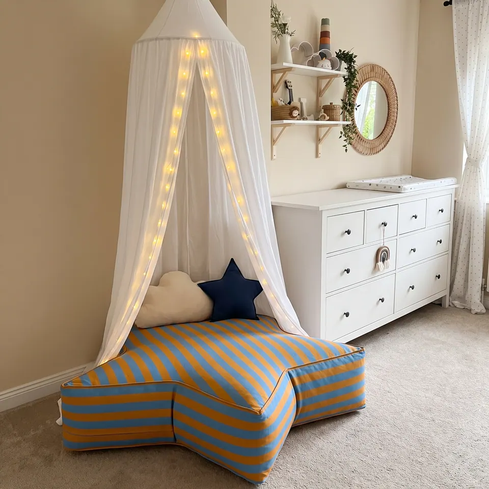 Blue and mustard awning-stripe star pouf under a fairy-light canopy beside a white changing dresser in a sunny nursery