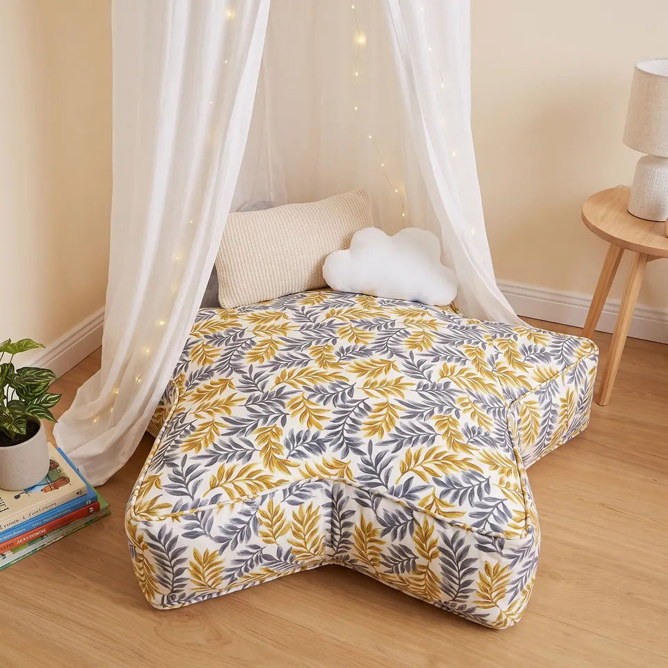 Leaf-print star floor cushion in cream, mustard and gray with a ribbed knit lumbar under a canopy beside an oak lamp table