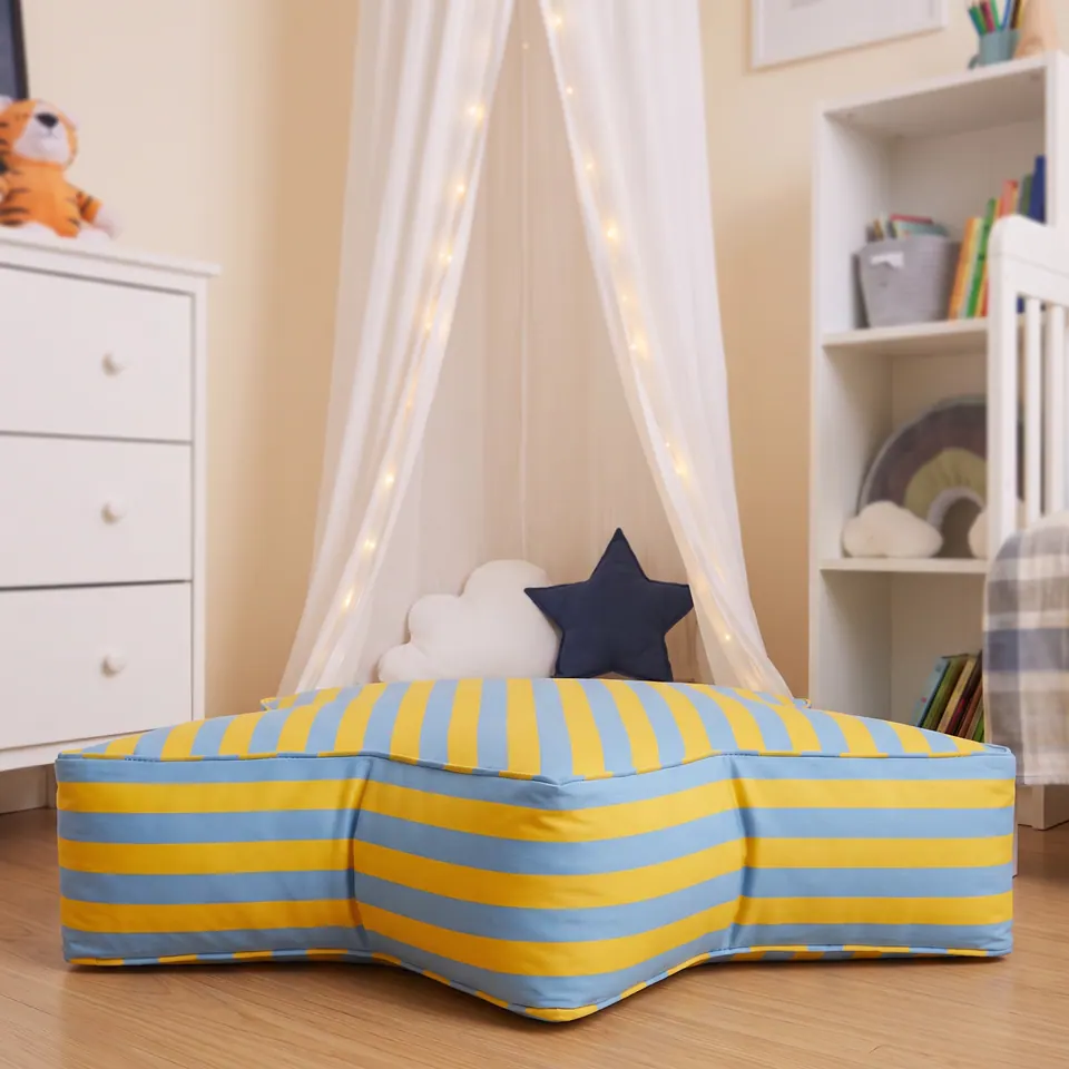 Front view of a yellow and powder blue striped star pouf at floor level with a navy star pillow, white dresser and bookcase