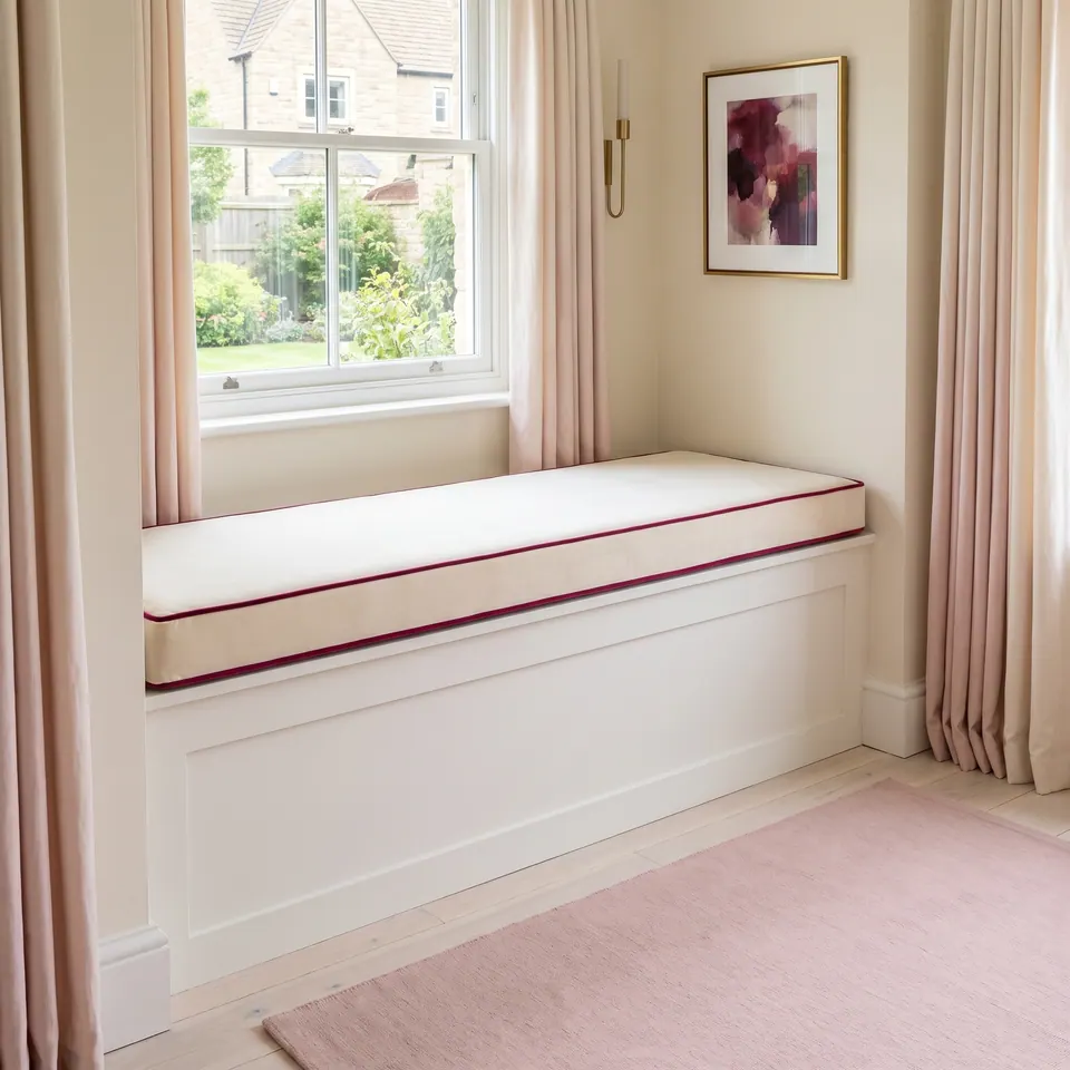 Cream velvet box cushion with fuchsia piping on a white paneled window seat between blush pink curtains
