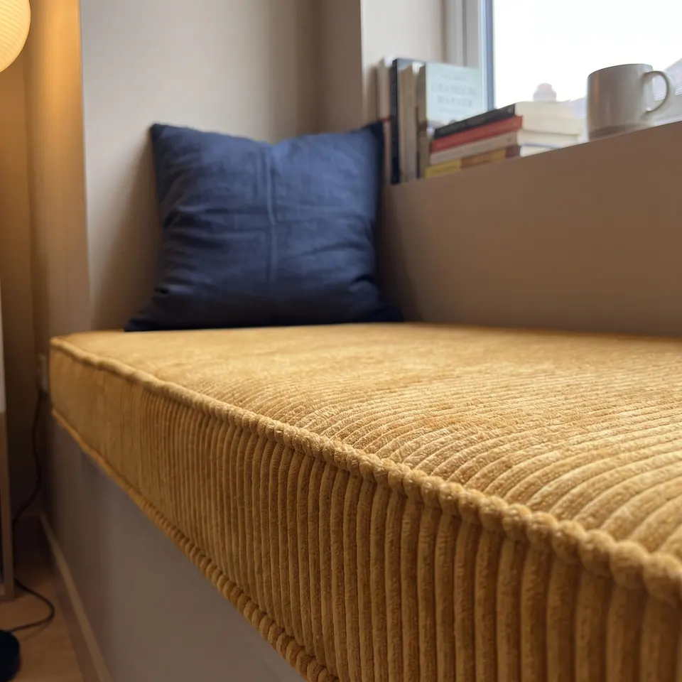 Close low-angle view of a mustard corduroy window seat cushion showing self piping and a ribbed gusset, navy pillow behind