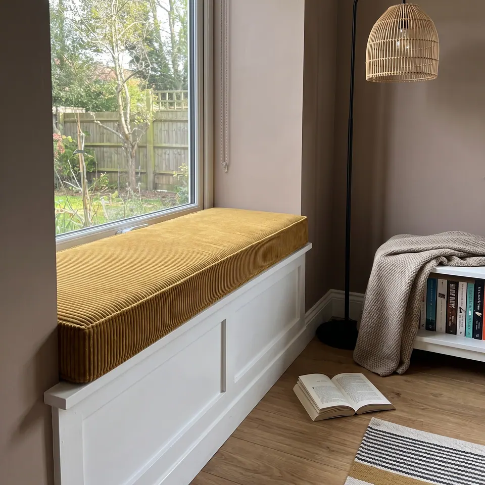 Mustard corduroy box cushion on a white paneled window seat, rattan floor lamp, knit throw and open book on the floor