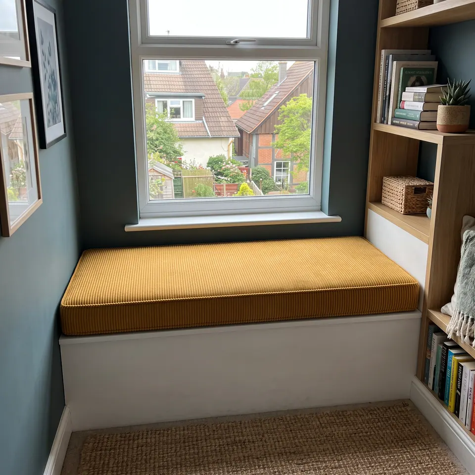 Mustard corduroy box cushion on a white window seat in a narrow teal nook with oak shelving and a rooftop view